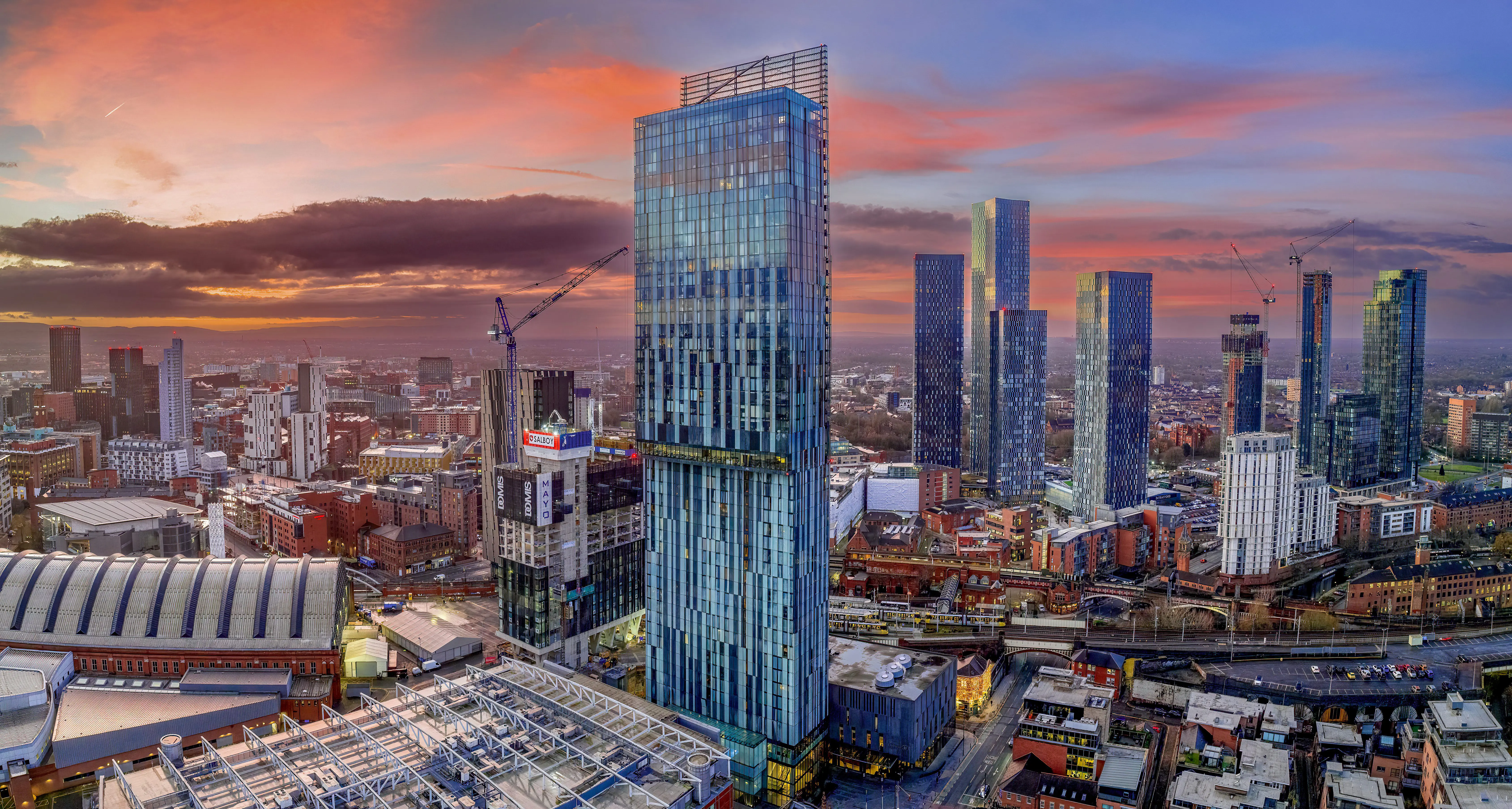 A city skyline at sunset with tall modern skyscrapers and cranes under a colorful sky.