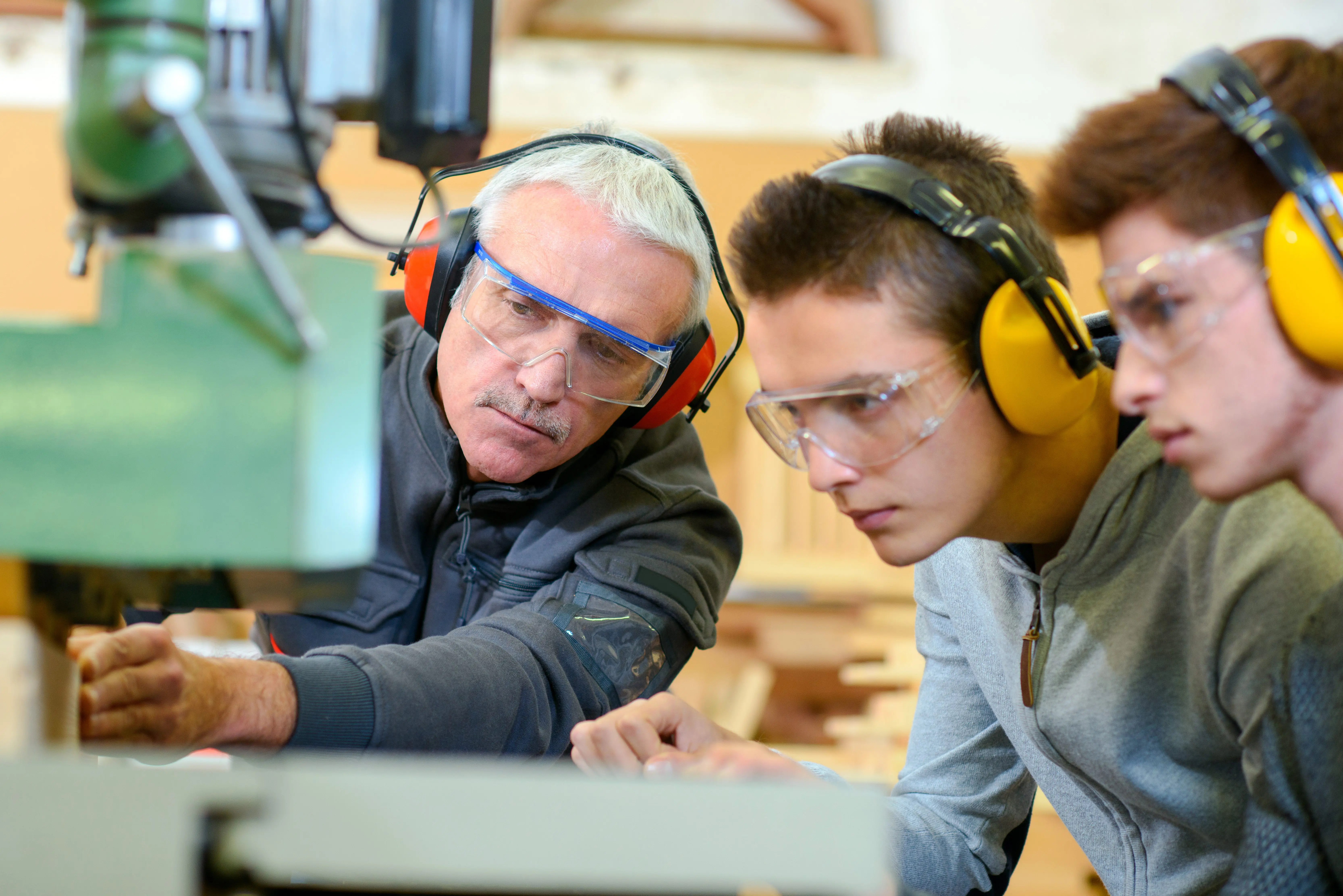 An instructor teaches two students woodworking, all wearing safety glasses and ear protection.