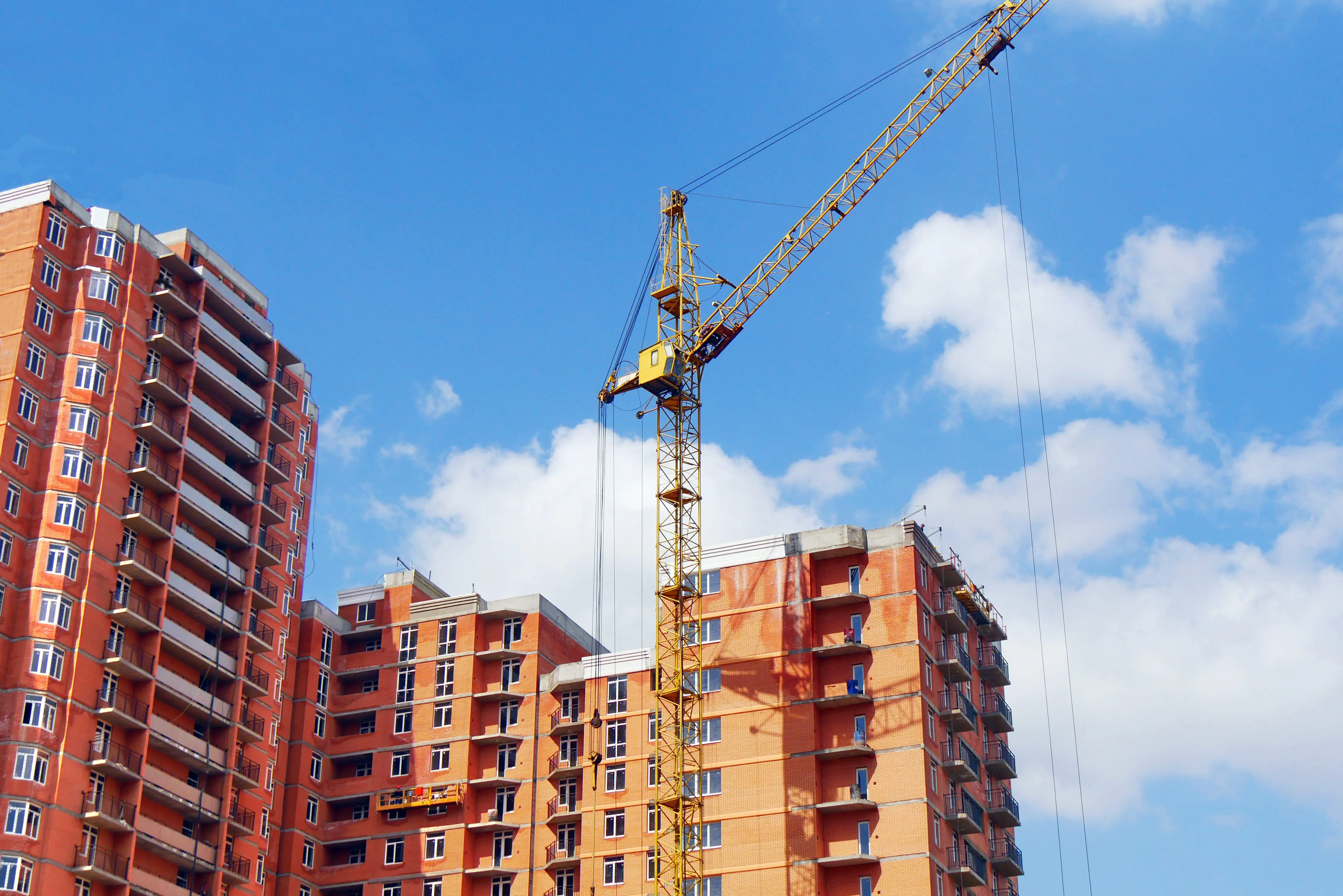 Yellow construction crane beside unfinished red brick apartments under a blue sky with scattered clouds.