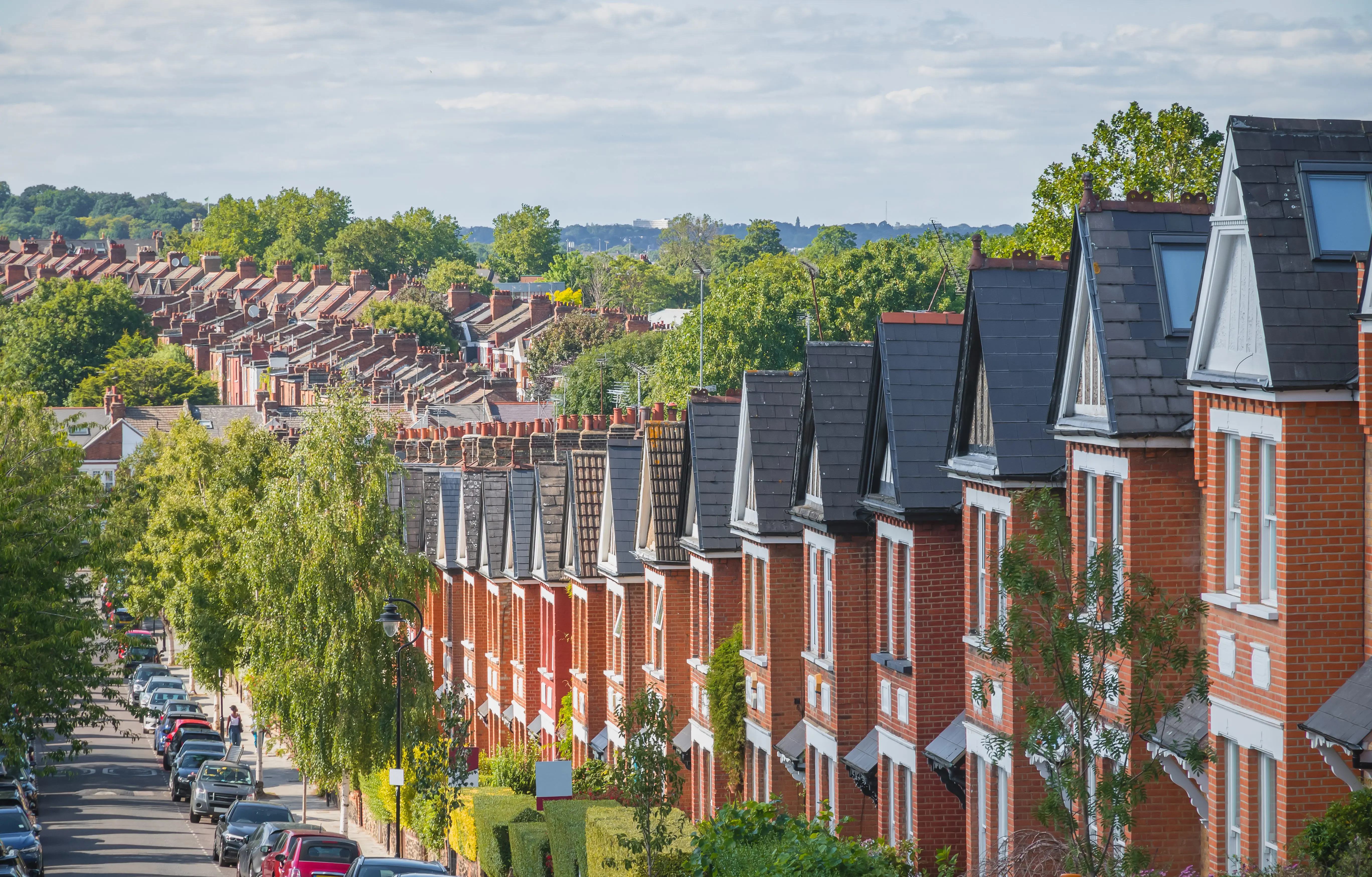 A row of red-brick terraced houses with parked cars and trees along a residential street.