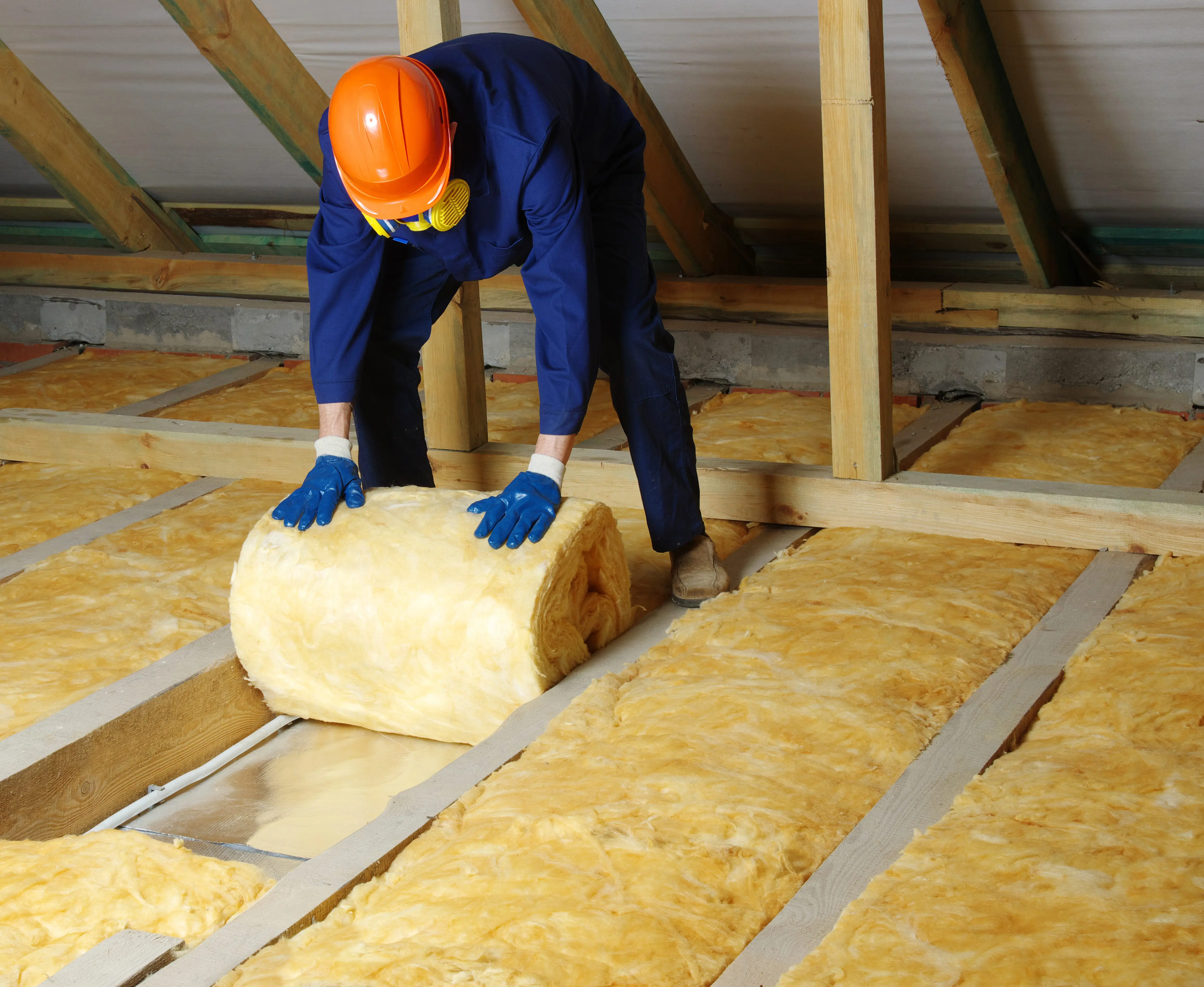 Worker in protective gear laying fiberglass insulation in an attic under a wooden roof structure.