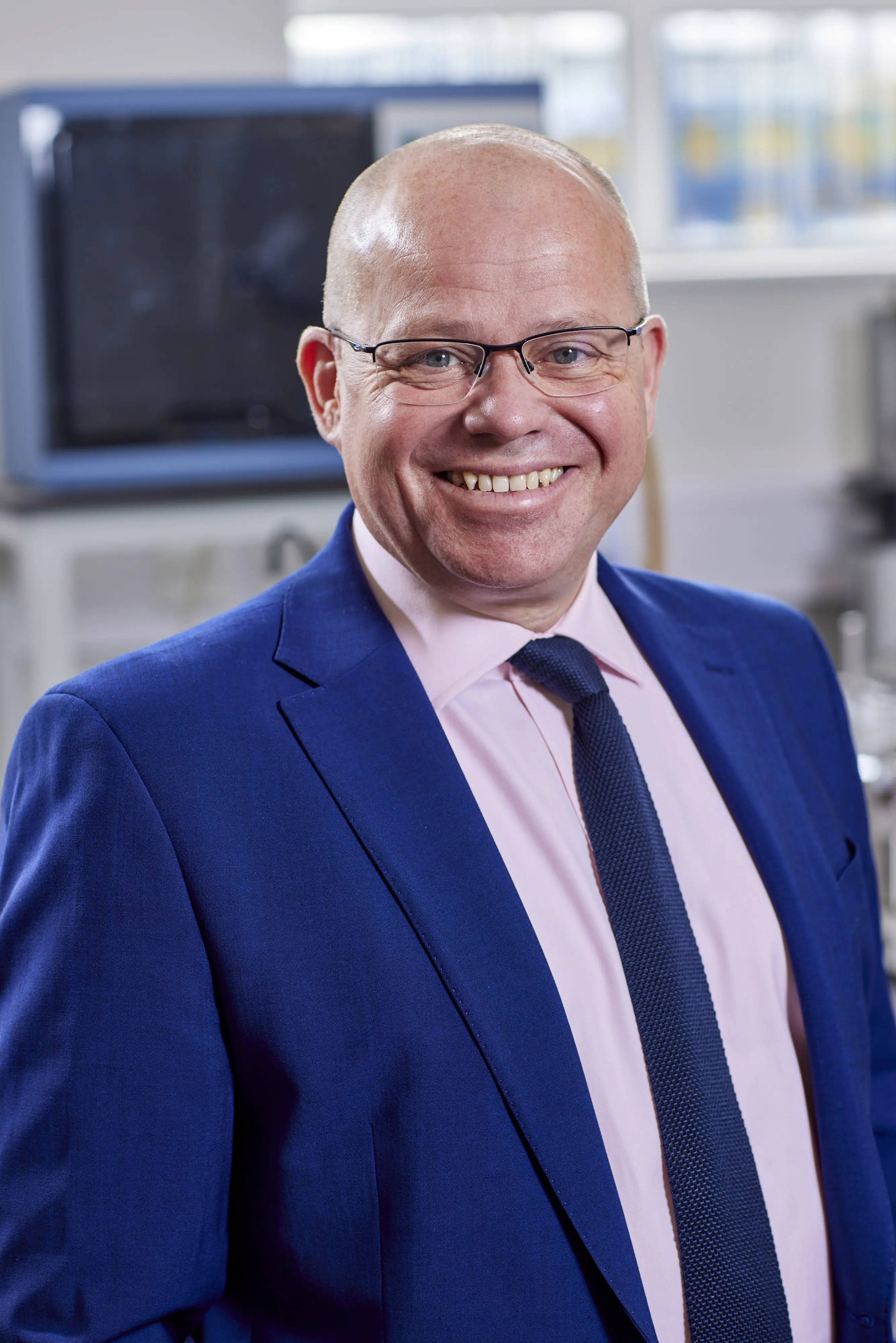 Smiling man in glasses, blue suit, pink shirt, and tie standing in an office or laboratory setting.