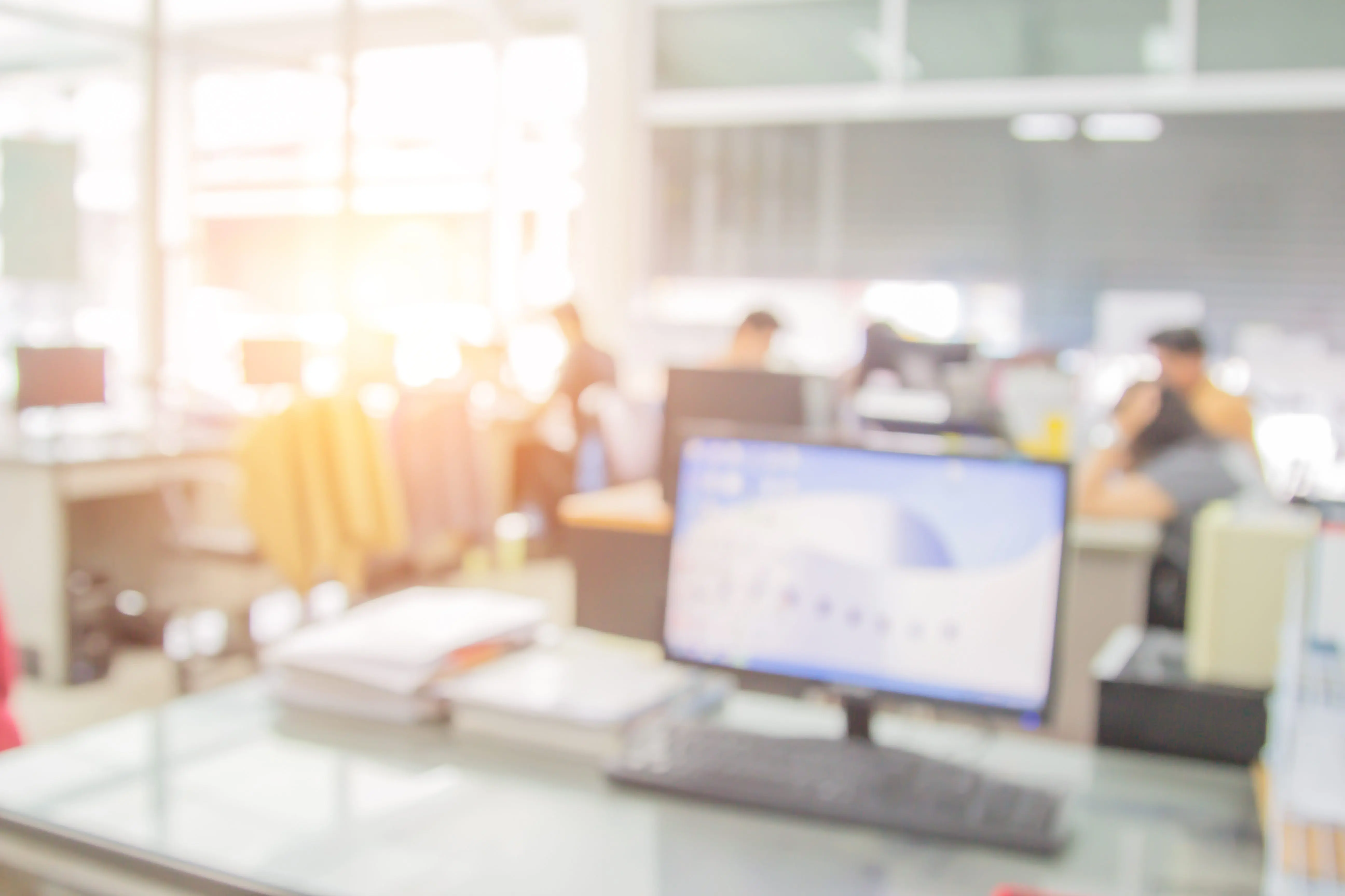 A blurry office scene with a computer on a desk and people working in the background.