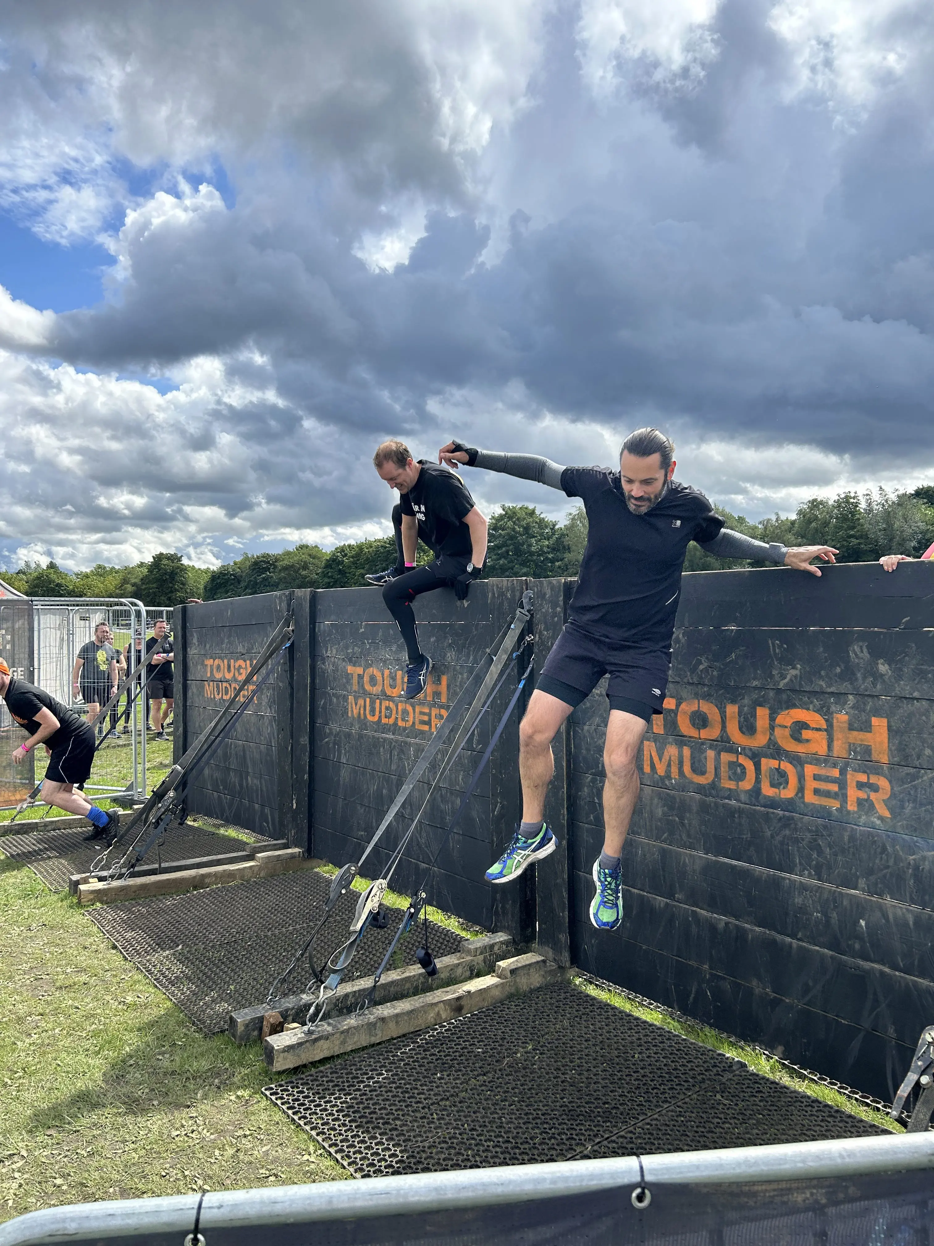 Two men in athletic gear jump over a Tough Mudder obstacle wall under a cloudy sky.