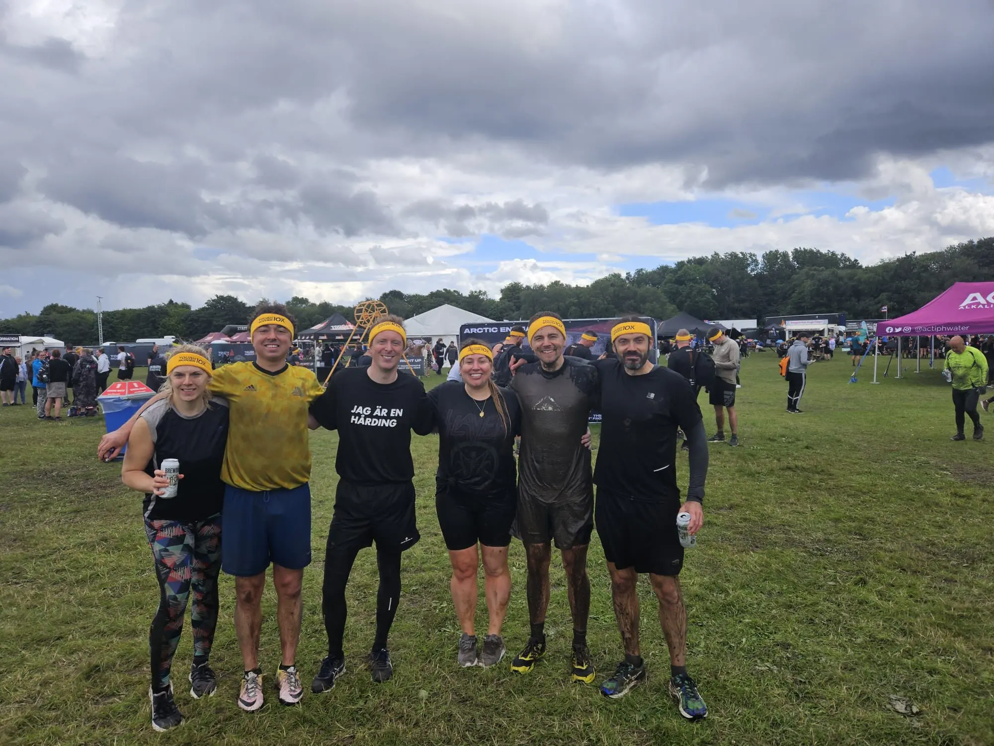 Six people in muddy athletic clothes and yellow headbands pose together outdoors after an obstacle race event.