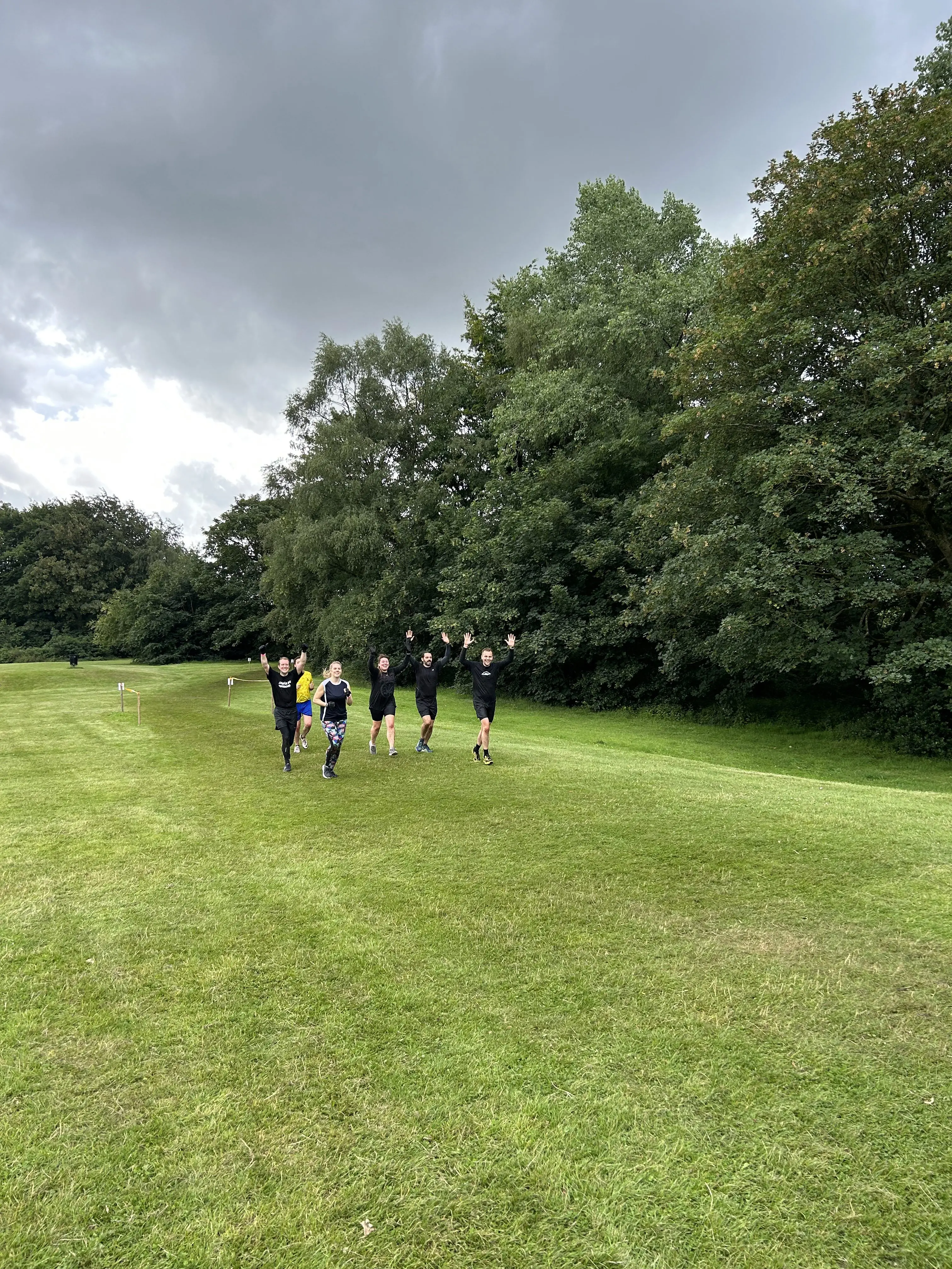 Six people run on a grassy field with their arms raised, surrounded by trees under a cloudy sky.