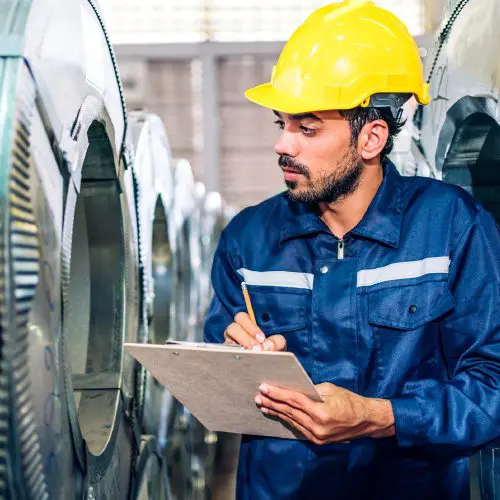 Man in a yellow hard hat and blue uniform inspects equipment and writes on a clipboard in a factory.