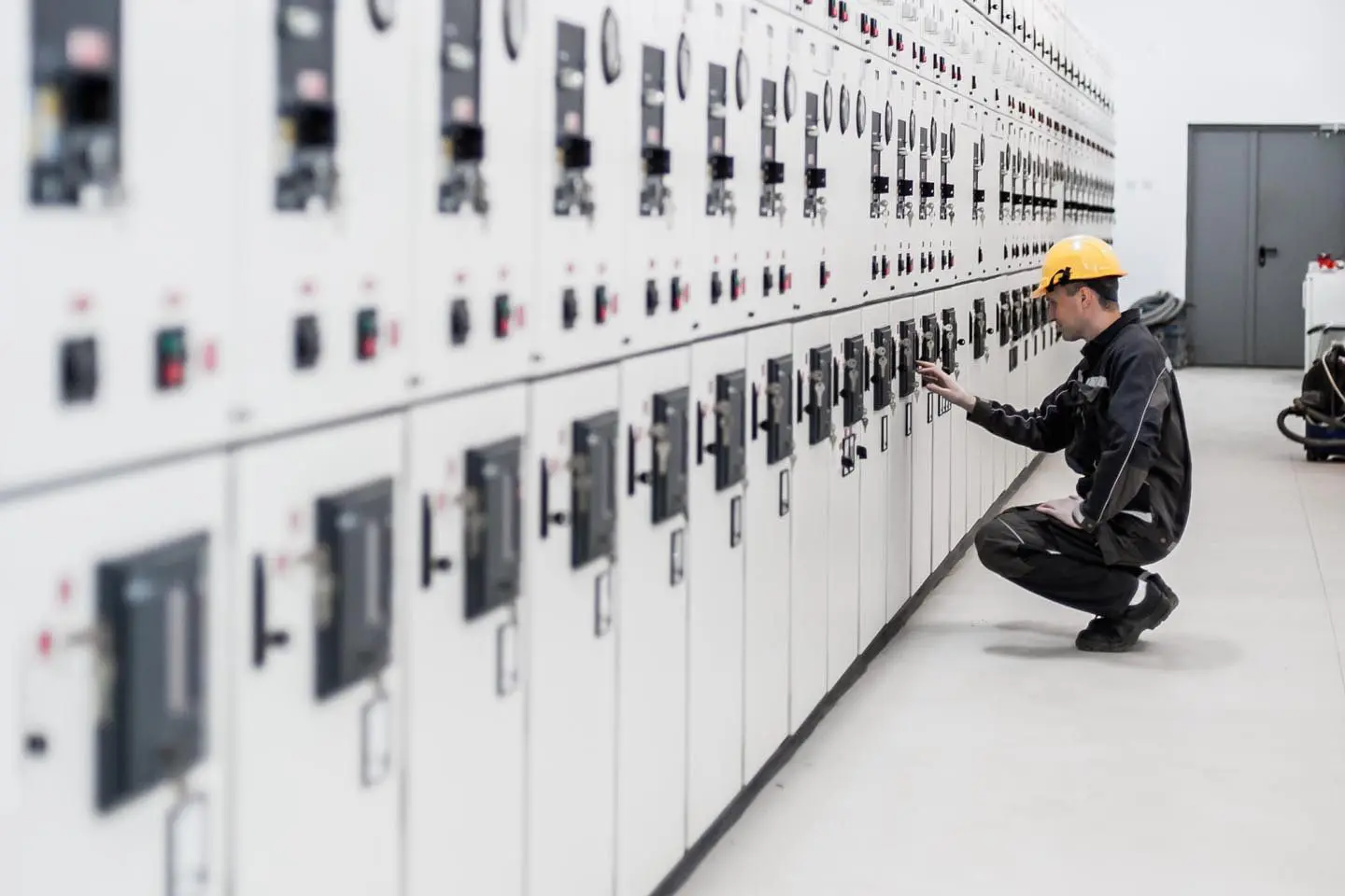 Worker in a hard hat operates control panels in an industrial electrical control room.