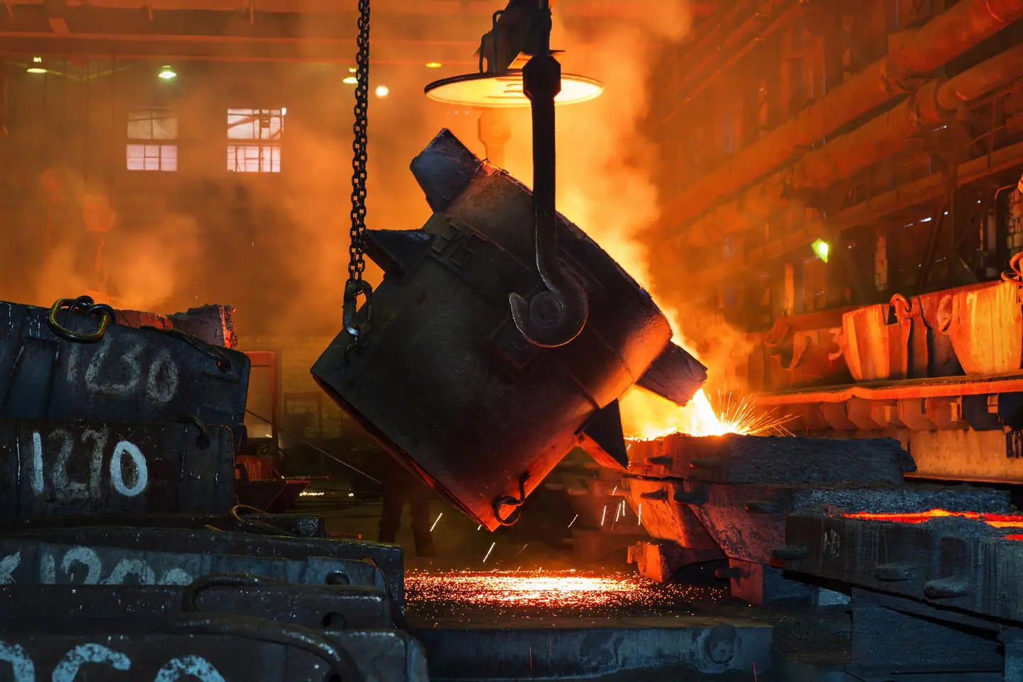 Molten metal pouring from a large ladle in a foundry with sparks and orange glow in the background.