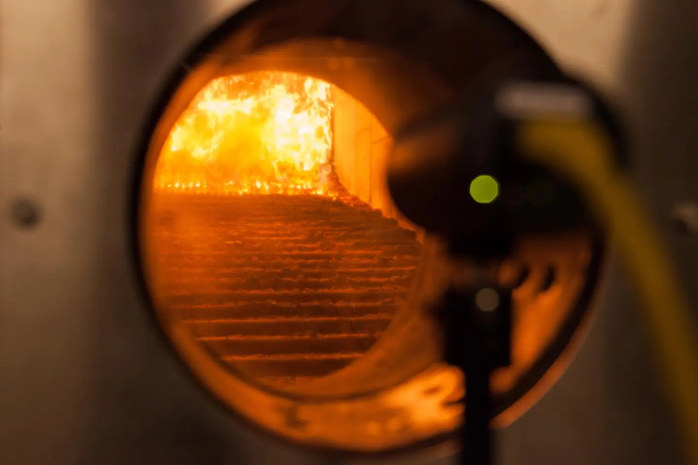 A view through a round window into a furnace with bright orange flames and metal steps inside.