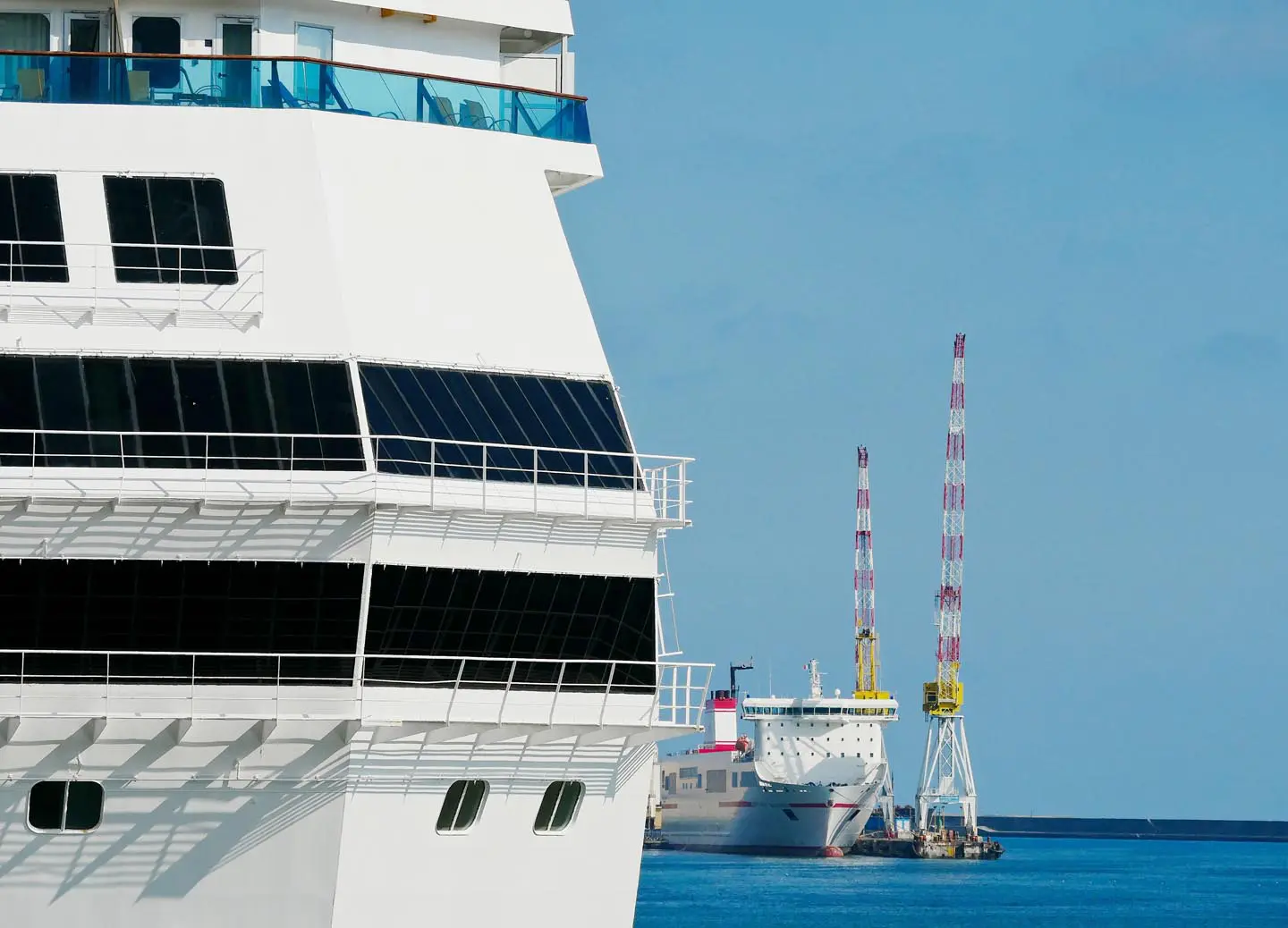 Close-up of a white cruise ship docked at a port, with another ship and cranes in the background.