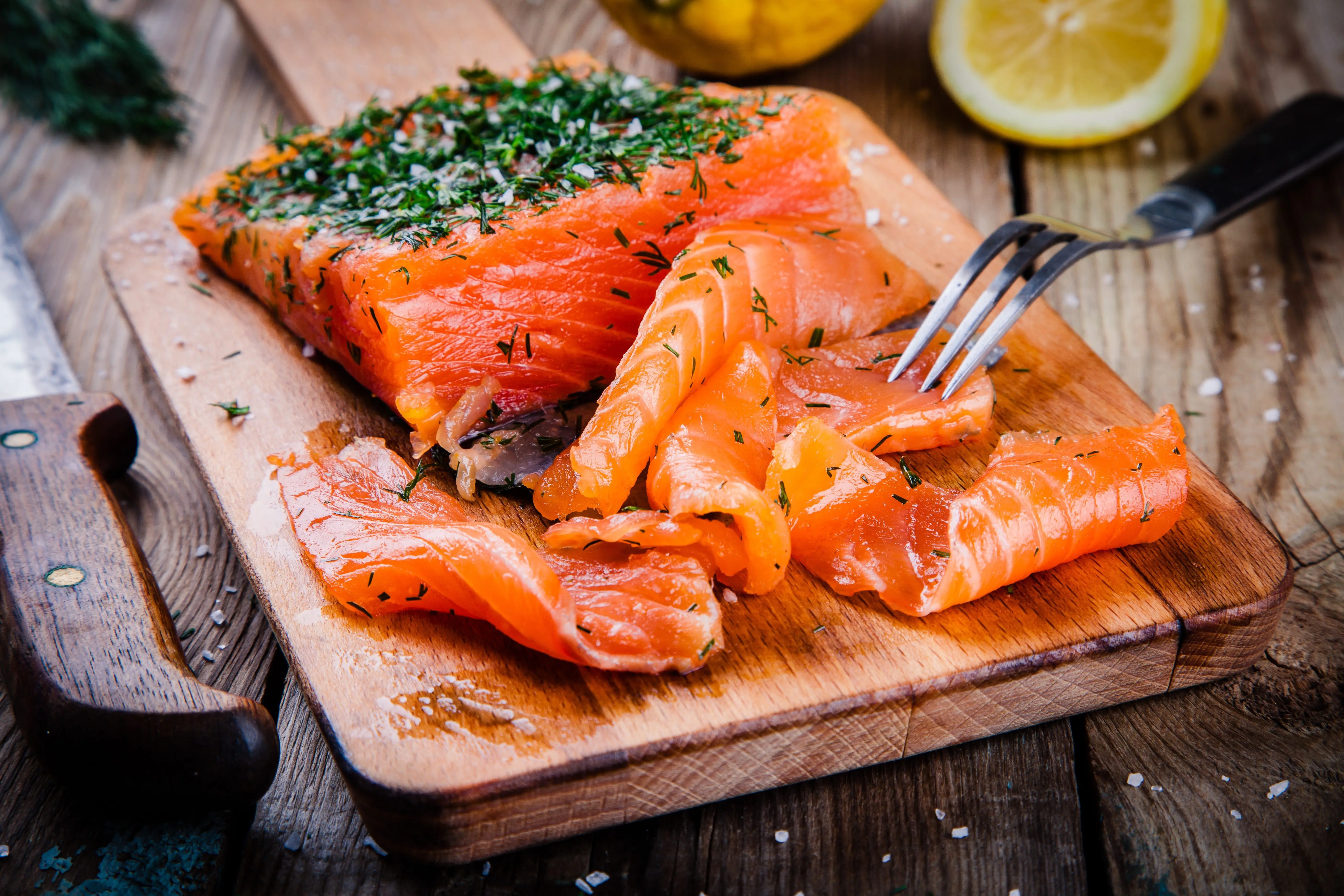 Sliced cured salmon with herbs on a wooden board, next to a fork, knife, and lemon halves.