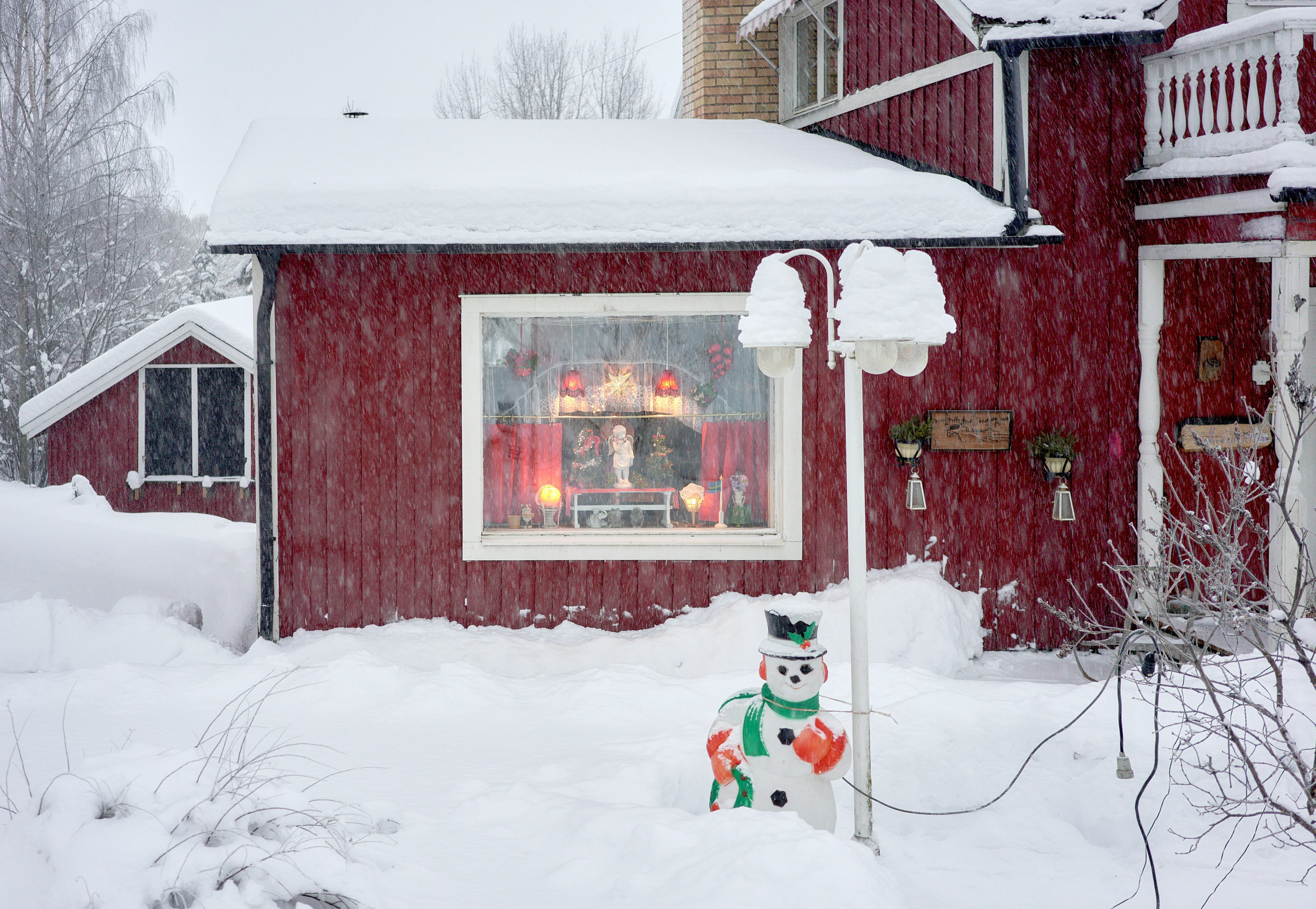 A snowman and a lamp post stand in deep snow outside a red house with a warmly lit window.