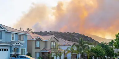 Smoke from a wildfire rises behind a suburban neighborhood with several houses and trees visible in the foreground.