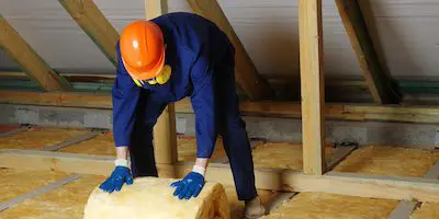 Worker in protective gear unrolling insulation material in an unfinished attic space.