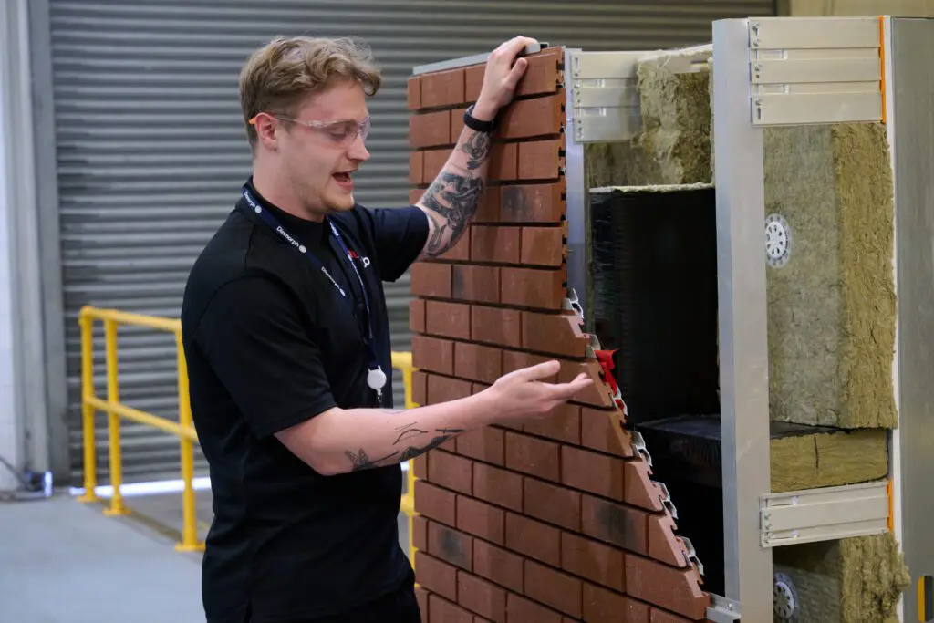 A man demonstrates attaching a brick cladding panel to an insulated wall in an industrial setting.