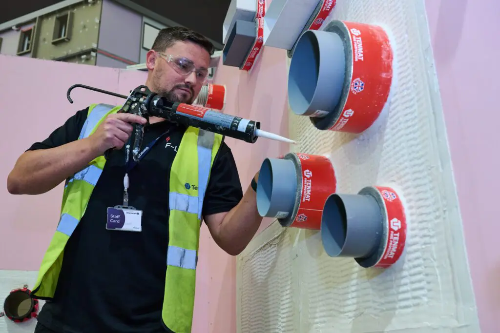 A man in safety gear applies sealant to grey pipes with red collars on a wall.