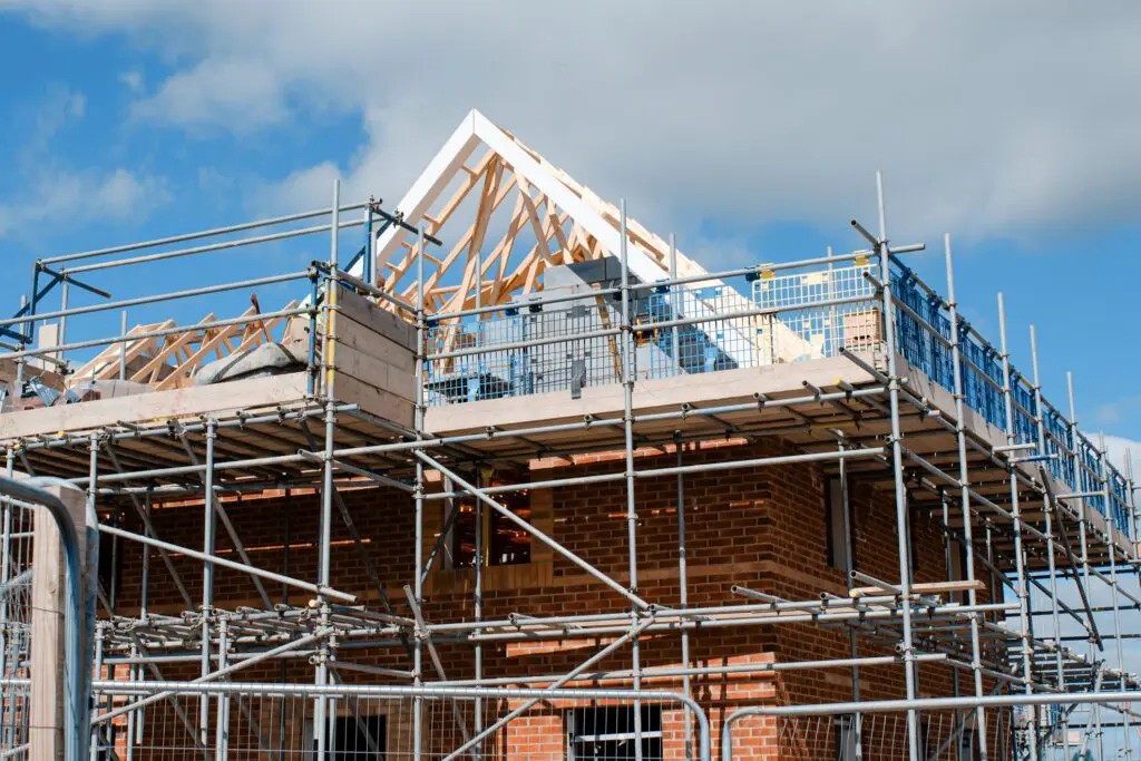 A brick house under construction with scaffolding and exposed wooden roof framing against a blue sky.