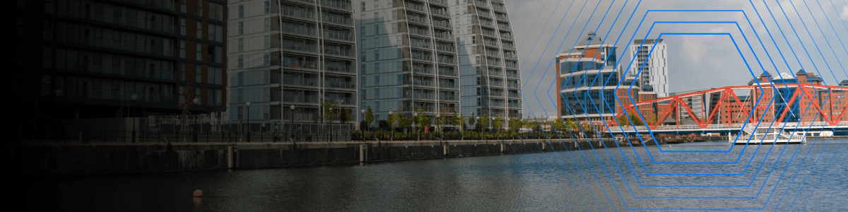 Modern waterfront buildings and a red bridge reflected in calm water under a partly cloudy sky.