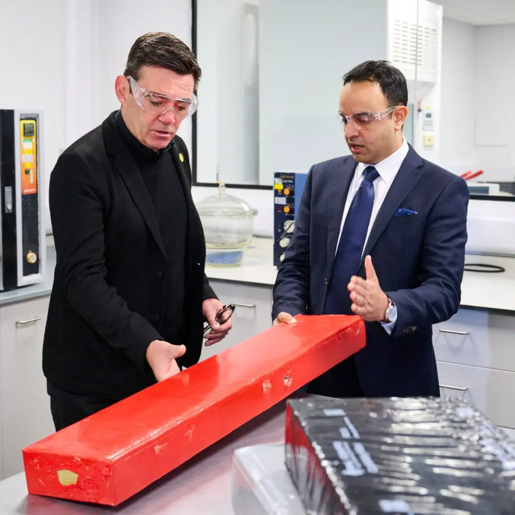 Two men in safety glasses examine a long red rectangular object in a laboratory setting.