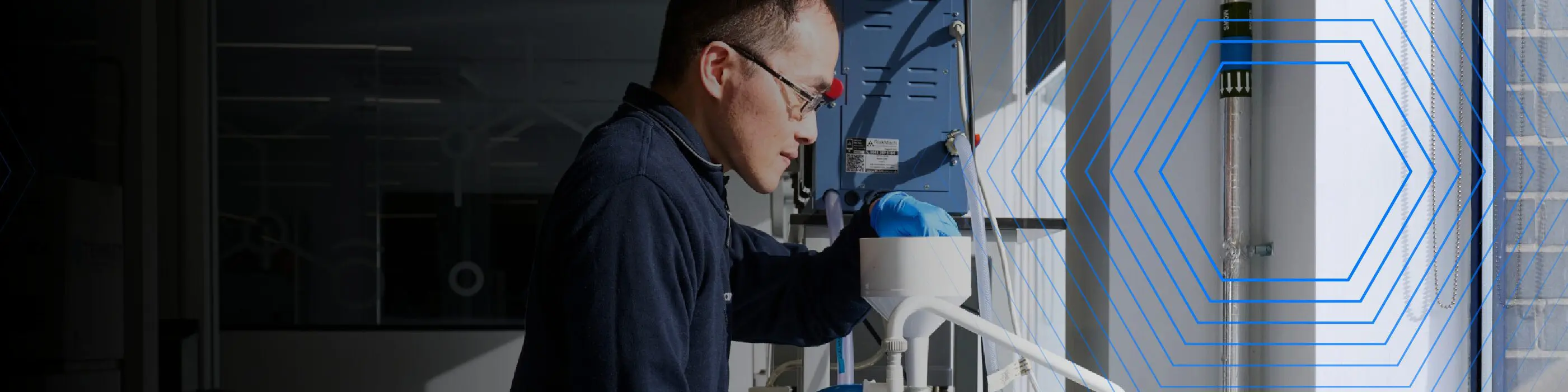 A person in safety gear operates laboratory equipment near a window with blue hexagonal graphics.