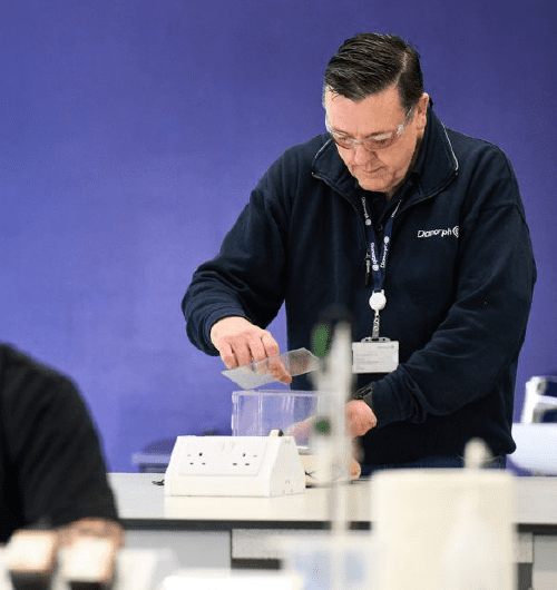 A man in a lab coat works with scientific equipment at a laboratory table against a purple background.