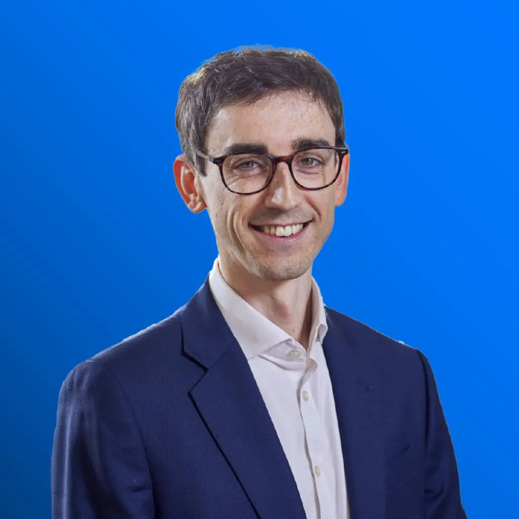 Man in glasses and a blue suit smiling, standing against a bright blue gradient background.