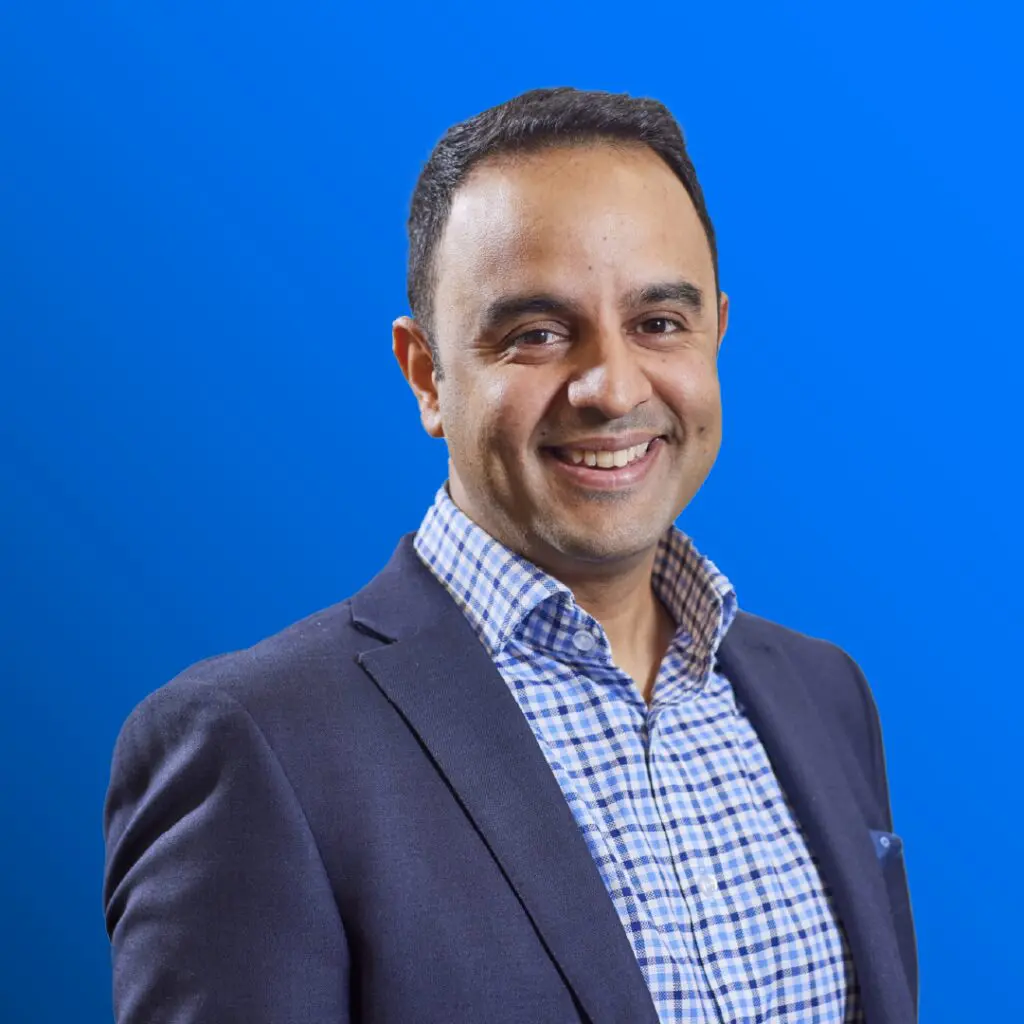 Man in a blue suit and checkered shirt smiling in front of a bright blue background.