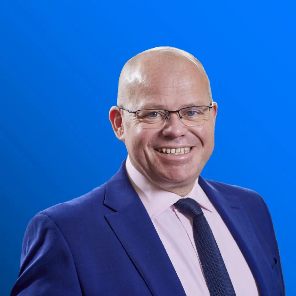 Smiling bald man in glasses, suit, and tie against a solid blue background.