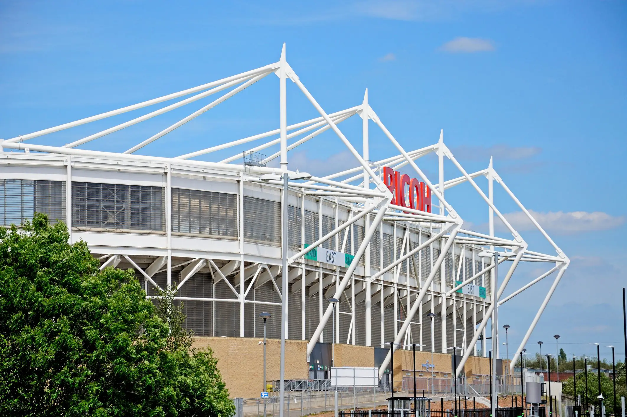 A modern stadium with white steel supports and a large SYON sign, under a blue sky with some clouds.