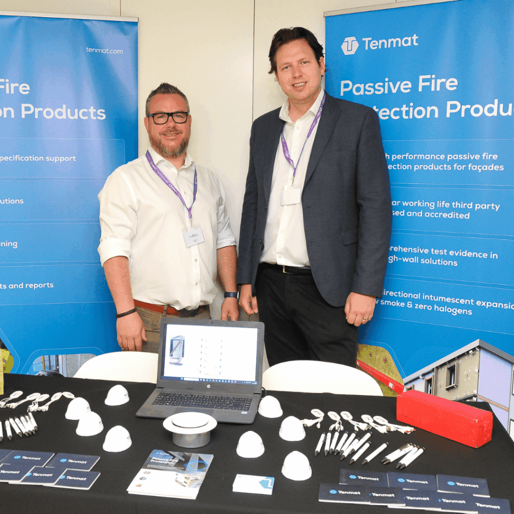 Two men stand at a Tenmat booth displaying passive fire protection products and informational materials.