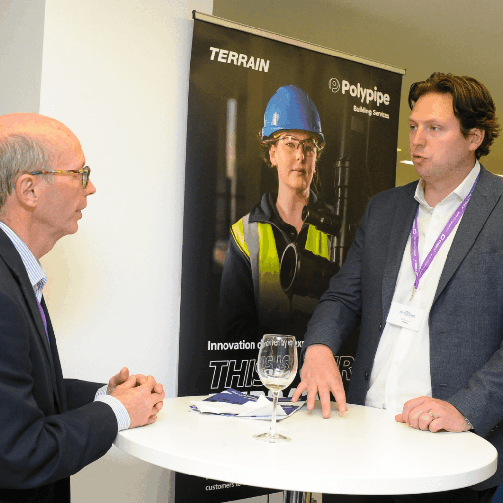 Two men talking at a round table in front of a Polypipe promotional banner at an event.