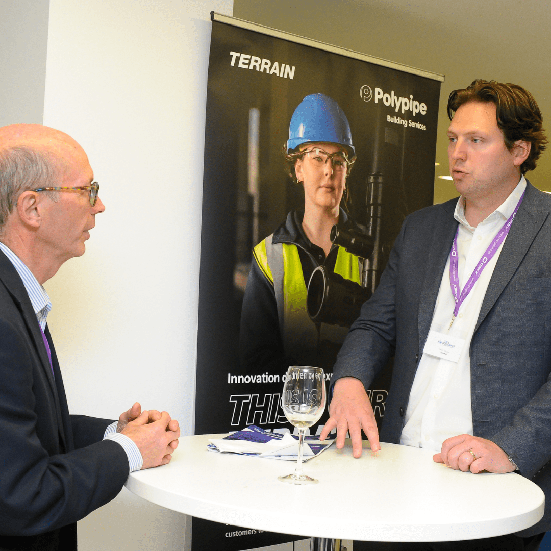 Two men talking at a round table in front of a Polypipe promotional banner at an event.