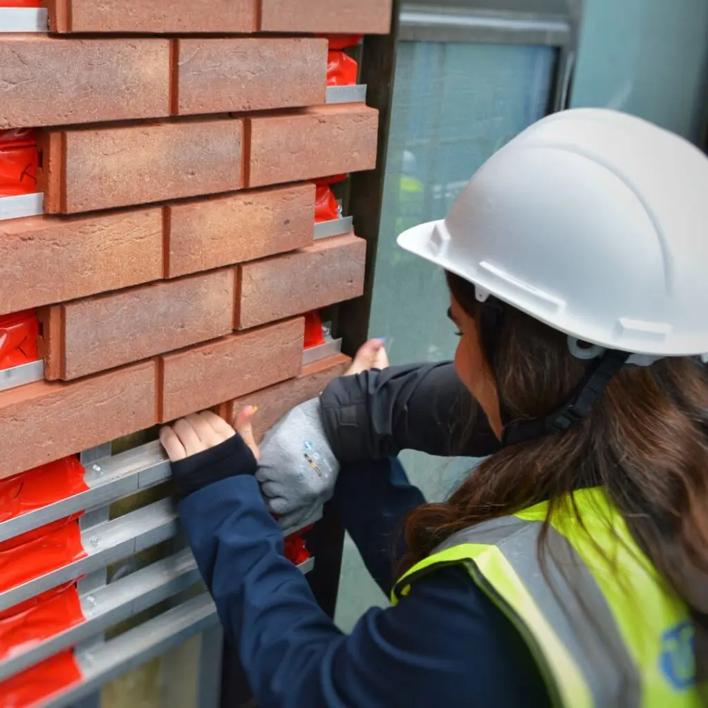 Person in a hard hat and vest installing brick facade panels on a wall at a construction site.
