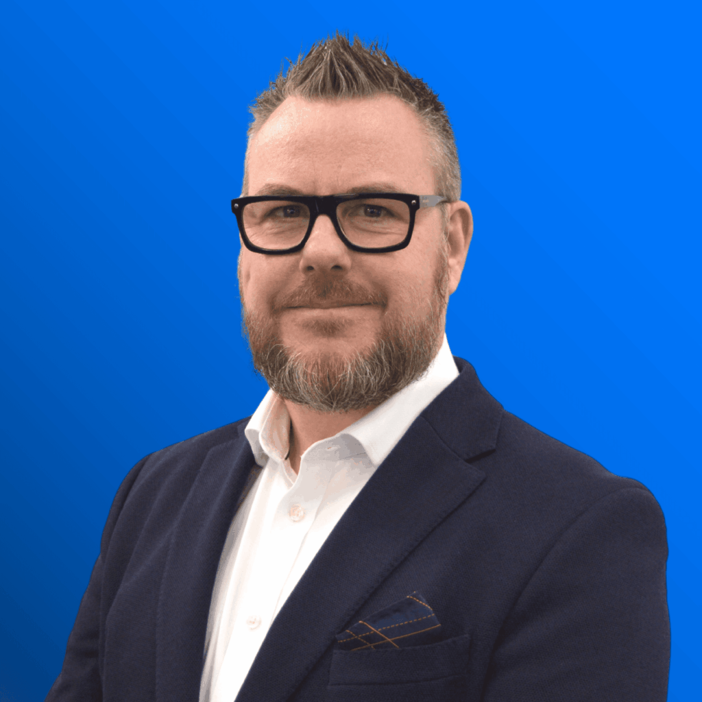 Man with beard and glasses wearing a navy suit and white shirt, posing against a blue background.