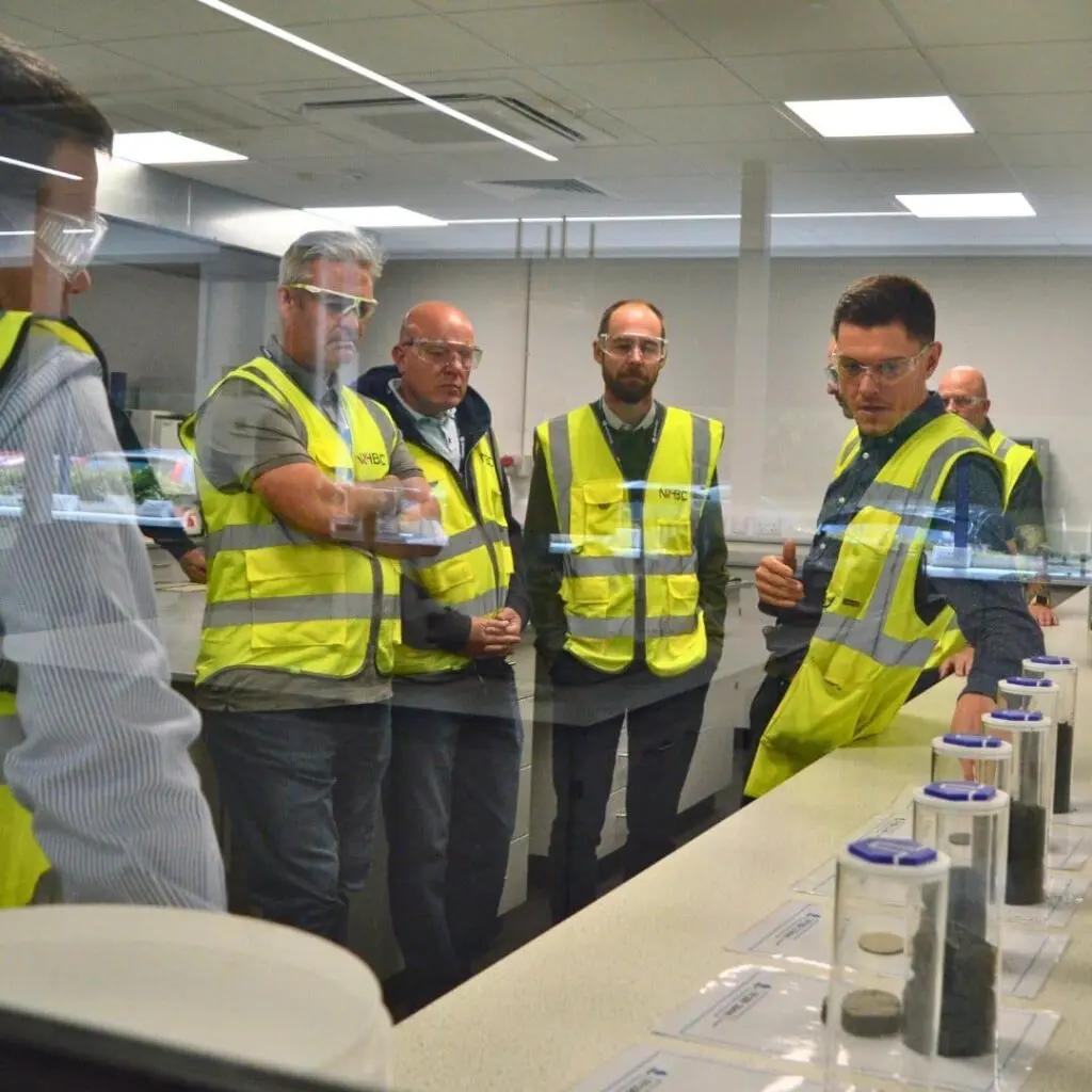 Five people in safety vests and goggles observe samples on a lab counter behind glass in a laboratory.