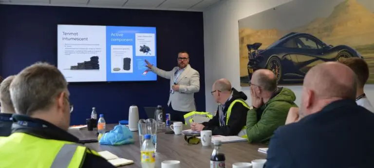 A man gives a presentation to a group seated at a table with a car photo and slideshow in the background.