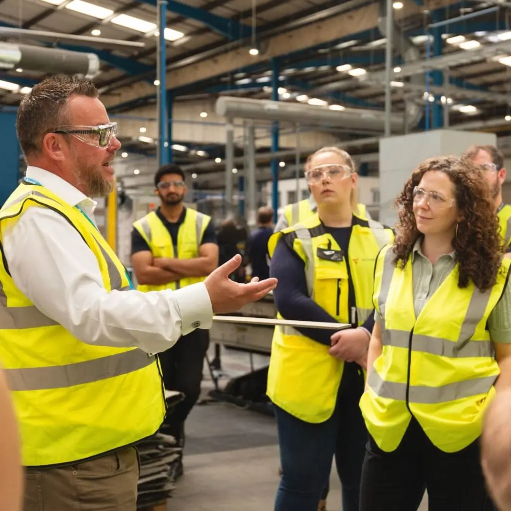 A man in a hi-vis vest speaks to a group of workers listening in a factory setting.