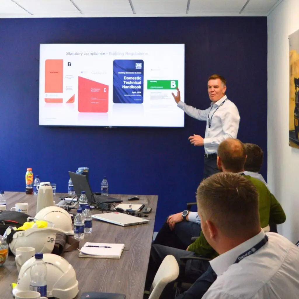 A man presents building regulations slides to colleagues in a conference room with hard hats on the table.