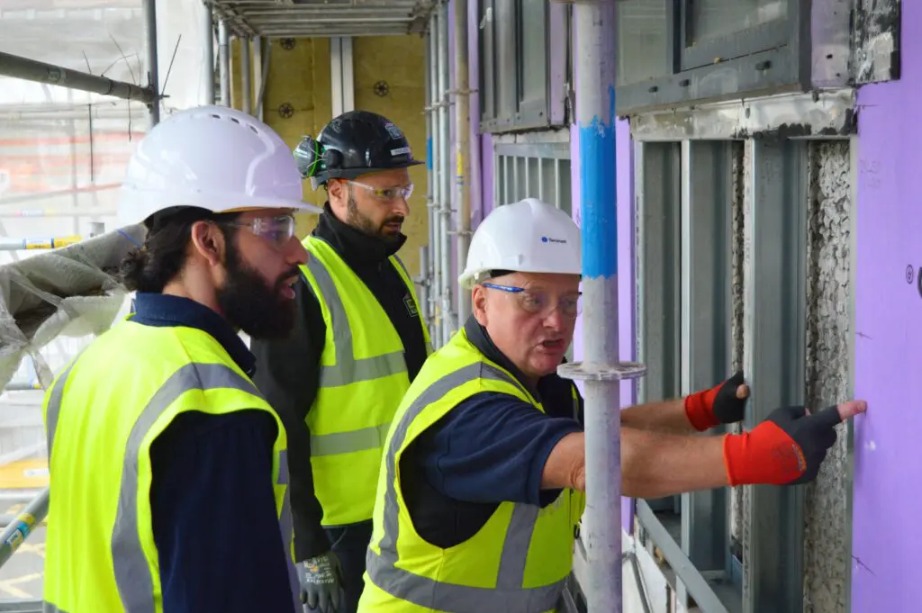 Three construction workers in safety gear talk by a purple wall at a building site.