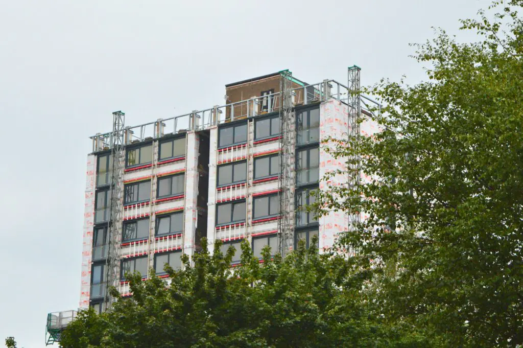 Tall building under construction with insulation panels, amid leafy trees.