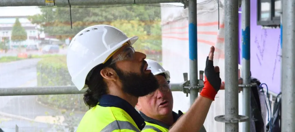 Two construction workers in hard hats inspect a building site; one points at a wall.