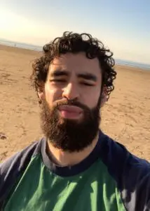 Curly-haired man with beard taking a beach selfie under a clear sky. Mohammad-Ali