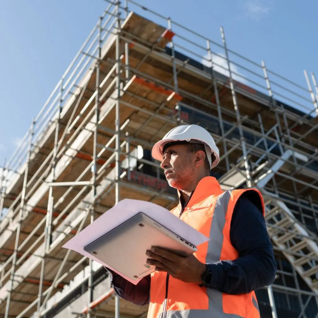 Man in safety vest and helmet holds documents before scaffolding on a clear day.