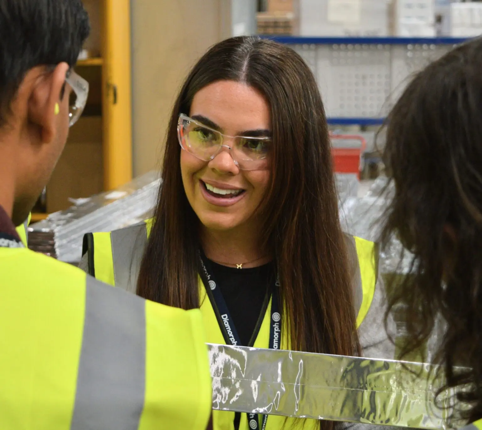 Woman in safety glasses and vest talks with colleagues in an industrial workplace.