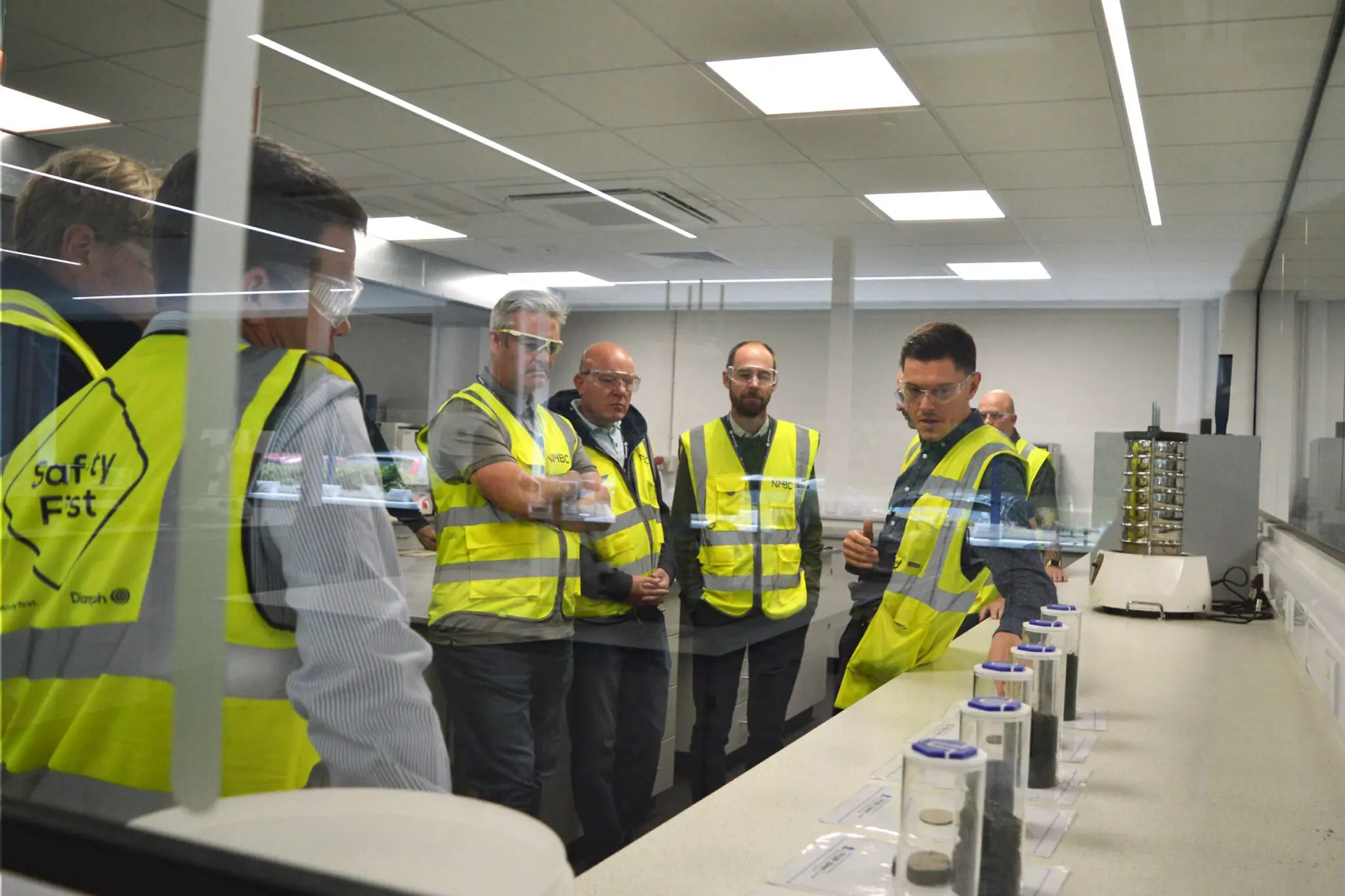 Six people in safety vests view fire protection samples on a lab counter behind glass.