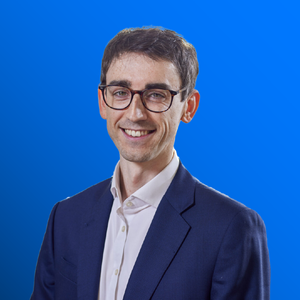 Smiling man in glasses and suit jacket against a blue gradient background.