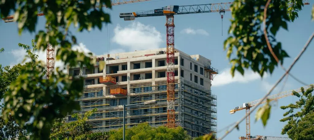 Construction site with cranes, surrounded by green trees beneath a blue sky.