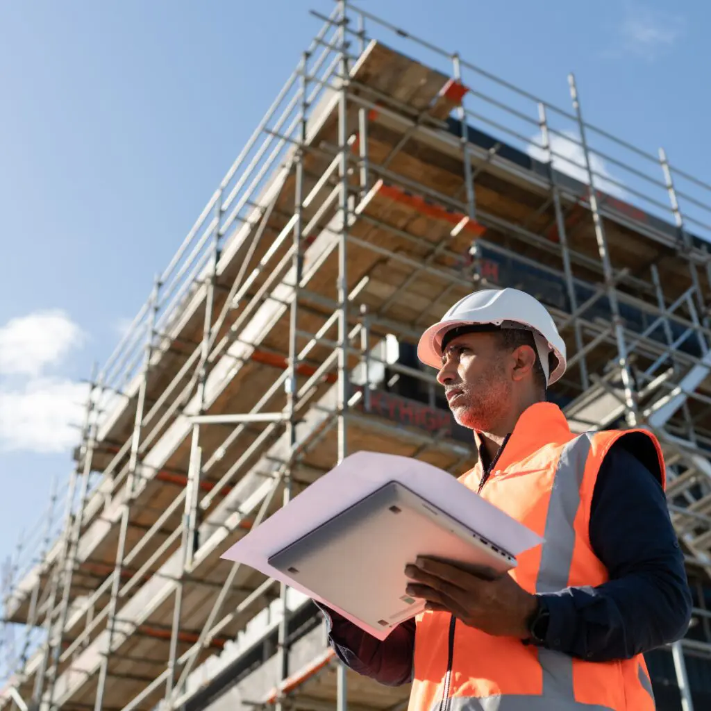 Worker in hard hat and vest holds papers beside scaffolding.