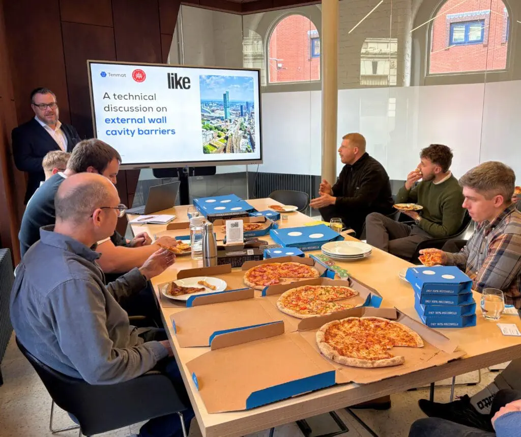 Six people eat pizza at a table during a technical talk on wall cavity barriers.